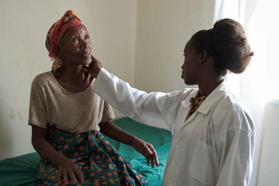 Foto de uma mulher médica, negra, vestindo jaleco branco, examina com toque abaixo da garganta outra mulher que está sentada na maca. A mulher usa turbante vermelho, roupa bege e olha para cima durante o exame. 
